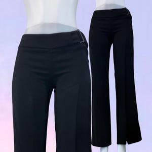 May include: A pair of solid black trousers featuring a high-waisted design with a side buckle detail on the waistband. The pants have a straight-leg cut with a clean, tailored appearance and a smooth fabric finish, displayed on a mannequin form.