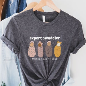 May include: A dark gray t-shirt with the text "expert swaddler" and a graphic of four swaddled babies. The text "mother baby nurse" is below the graphic.