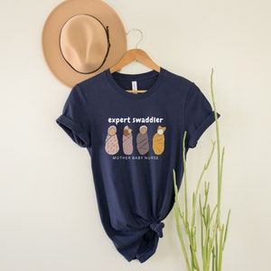 May include: Navy blue t-shirt with a white graphic of four swaddled babies and the text "expert swaddler" and "mother baby nurse".