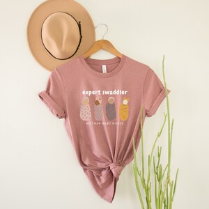 May include: A pink t-shirt with the text "expert swaddler" and a graphic of three swaddled babies. The text "mother baby nurse" is below the graphic.