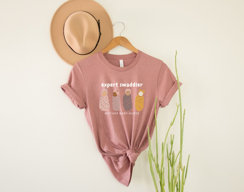 May include: A pink t-shirt with the text "expert swaddler" and a graphic of three swaddled babies. The text "mother baby nurse" is below the graphic.