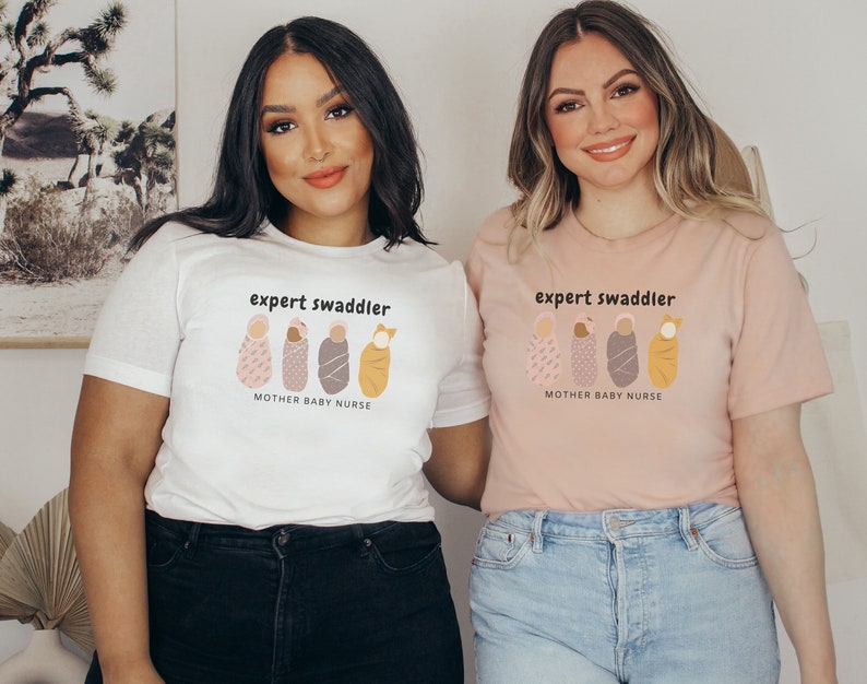May include: Two people wearing t-shirts with the text "expert swaddler" and a graphic of four swaddled babies. The text below the graphic reads "mother baby nurse". One shirt is white and the other is pink.