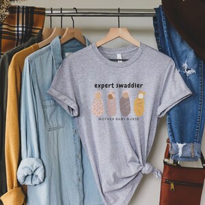 May include: Gray t-shirt with the text "expert swaddler" and a graphic of four swaddled babies. The text below the graphic reads "mother baby nurse".