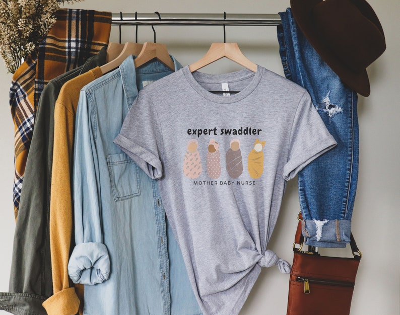 May include: Gray t-shirt with the text "expert swaddler" and a graphic of four swaddled babies. The text below the graphic reads "mother baby nurse".