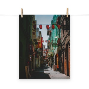 May include: A narrow street in Chinatown with red lanterns hanging overhead. The street is lined with buildings, some of which have Chinese characters on their signs. The street is empty except for a few people walking.