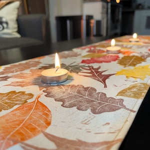 May include: A white table runner with a fall leaf design and three lit tea lights. The runner is on a dark wood table.