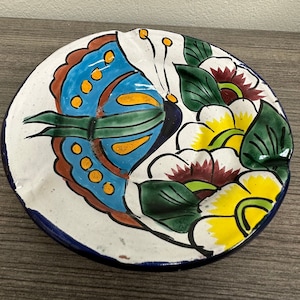 May include: A white ceramic plate with a blue, yellow, and green butterfly design. The butterfly is surrounded by white flowers with yellow centers and green leaves. The plate has a blue rim.