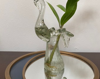 Op de afbeelding: Een heldere glazen vaas in de vorm van een zwaan met een groene plant erin. De vaas staat op een ronde, goudomrande spiegel met een blauw centrum.