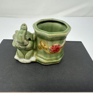 Könnte beinhalten: Ein grüner Keramik-Pflanztopf in Form eines Elefanten mit floralen Details. Der Topf hat eine große Öffnung zum Pflanzen.