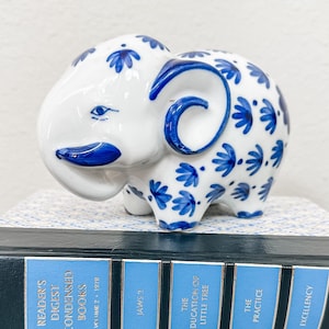 May include: A white ceramic elephant figurine with blue and white floral patterns. The elephant is sitting on top of a stack of blue and white books. The books have titles like "Jaws 2" and "The Education of Little Tree".