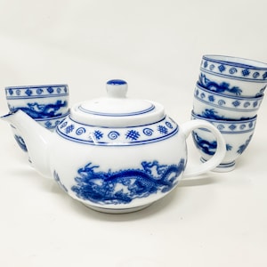 Op de afbeelding: Witte porseleinen theeset met blauwe draken- en wervelpatronen. De set bestaat uit een theepot, twee theekopjes en een stapel van vier theekopjes. De theepot heeft een deksel en een handvat. De kopjes hebben een ronde vorm.