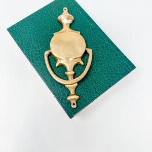 May include: A brass door knocker with a circular design and a decorative handle. The knocker is sitting on a green book.