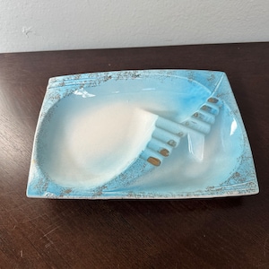 Könnte beinhalten: Ein hellblauer Aschenbecher aus Keramik mit weißem Innenraum und goldenen Akzenten. Der Aschenbecher hat ein einzigartiges Design mit einem erhöhten Bereich für Zigaretten.