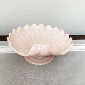 May include: A pink glass bowl with a scalloped edge and a pedestal base. The bowl is shaped like a seashell.
