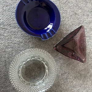 May include: Three different ashtrays. One is a dark blue ceramic ashtray with a rounded shape. Another is a clear glass ashtray with a diamond pattern. The third is a purple glass ashtray with a triangular shape.