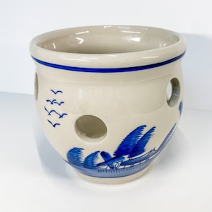 May include: A white ceramic onion keeper with blue and white painted designs. The keeper has a round shape with holes around the sides and a blue rim.