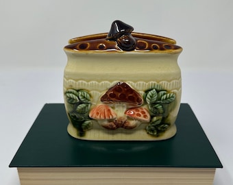 May include: A ceramic napkin holder shaped like a mushroom with brown and green details. The holder has a mushroom design on the front and a small mushroom on the top.