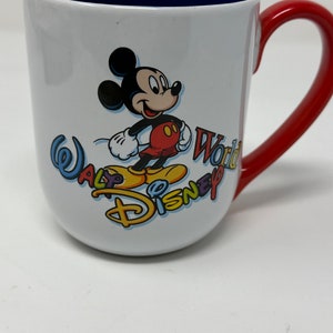 May include: White ceramic mug with a red handle. The mug features a colourful Walt Disney World logo with Mickey Mouse in the centre.