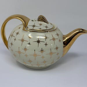 Könnte beinhalten: Eine weiße Teekanne mit einem goldenen Sternenmuster und goldenen Verzierungen. Die Teekanne hat einen runden Korpus und einen geschwungenen Ausguss.