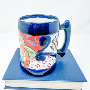 May include: A blue ceramic mug with a colourful floral design sits on top of a blue book. The mug has a white interior and a handle.