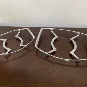 May include: Two silver metal hot pads with a wavy design. The hot pads are oval shaped and have four legs.