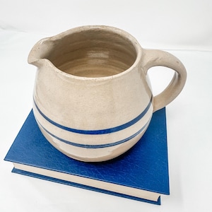 Könnte beinhalten: Ein cremefarbener Keramik-Krug mit zwei blauen Bändern um die Mitte. Der Krug hat einen runden Körper und einen Henkel. Er steht auf einem blauen Buch.