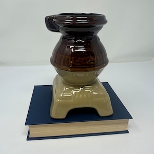 Könnte beinhalten: Eine Keramik-Ofen-Tasse in Form eines Holzofens. Die Tasse ist braun und beige mit einem Griff an der Seite. Die Tasse steht auf einem blauen Buch.