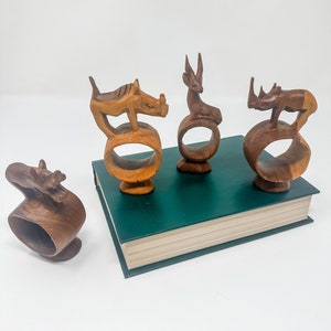 May include: Four wooden napkin rings shaped like animals. The rings are made of dark brown wood and feature a rhinoceros, a deer, a rhinoceros, and a stylized animal. The rings are sitting on a green book.