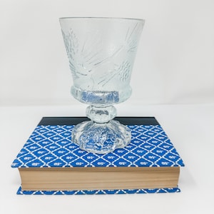 May include: A clear glass goblet with a pine cone design sits on top of a blue and white patterned book.
