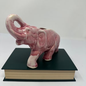 May include: A pink ceramic elephant planter with its trunk raised in the air. The planter is sitting on a dark green book.