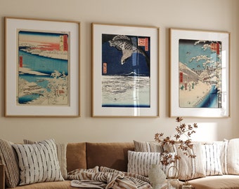 May include: A brown leather sofa with three throw pillows and a throw blanket. The sofa is in front of a wall with three framed Japanese woodblock prints. A wooden coffee table with a woven design sits in front of the sofa.