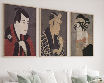 May include: Three framed Japanese woodblock prints depicting portraits of people in traditional clothing. The prints are in black, white, red, and gold tones and are displayed on a white wall.