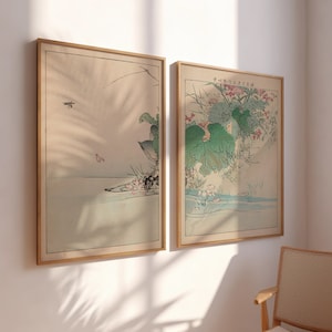 May include: Two framed prints of Japanese art. The print on the left shows a bird flying over a body of water with a large leaf in the background. The print on the right shows a large green leaf with pink flowers and a body of water in the background. The prints have a light brown frame.