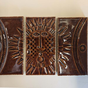 May include: Three brown ceramic tiles featuring a raised design of a stylised sun face. The sun face has a chequered pattern on the forehead and a circle around the eyes. The tiles are arranged horizontally.