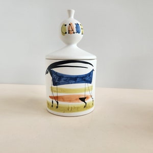 May include: White ceramic jar with a lid featuring a stylised animal design in black, blue, yellow, and orange. The lid has the word "CAFE" written in colourful letters.