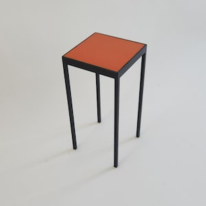 May include: A modern side table with a square, orange top and a black metal frame. The table has four slender legs and stands approximately 24 inches tall.