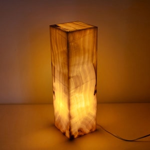 May include: A rectangular, translucent, white onyx lamp with a warm, yellow glow. The lamp is lit and has a white cord.