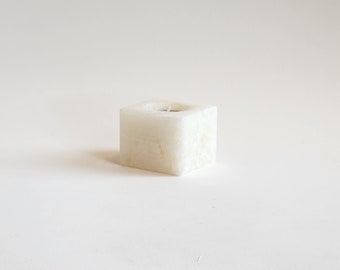 May include: A white square candle holder made of alabaster. The holder has a small, round opening in the center for a tea light candle.