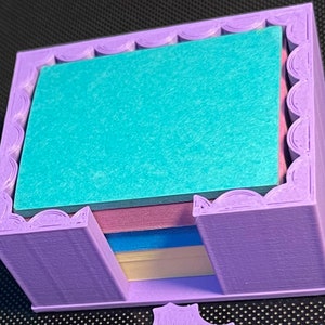 May include: A purple 3D printed holder for sticky notes. The holder has a decorative scalloped edge and is designed to hold multiple stacks of notes. The holder is empty, but there are four stacks of notes visible in the background.