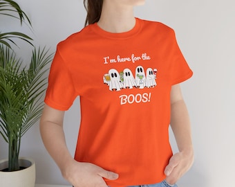 May include: A bright orange short-sleeve t-shirt featuring a graphic of four white ghosts holding various alcoholic drinks. Above the ghosts is the white text I'm here for the and below them is the white text BOOS! in a bold, playful font.
