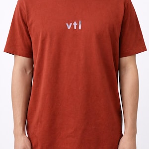 May include: Rust-colored short-sleeved t-shirt with the text "vtl" in a reflective, iridescent font across the chest. The shirt is made of a soft material and has a classic crew neck.