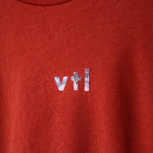 May include: Close-up of a rust-colored t-shirt with a crew neck. The shirt features the letters "vtl" in a light blue and white floral pattern on the chest. The fabric has a slightly textured appearance.