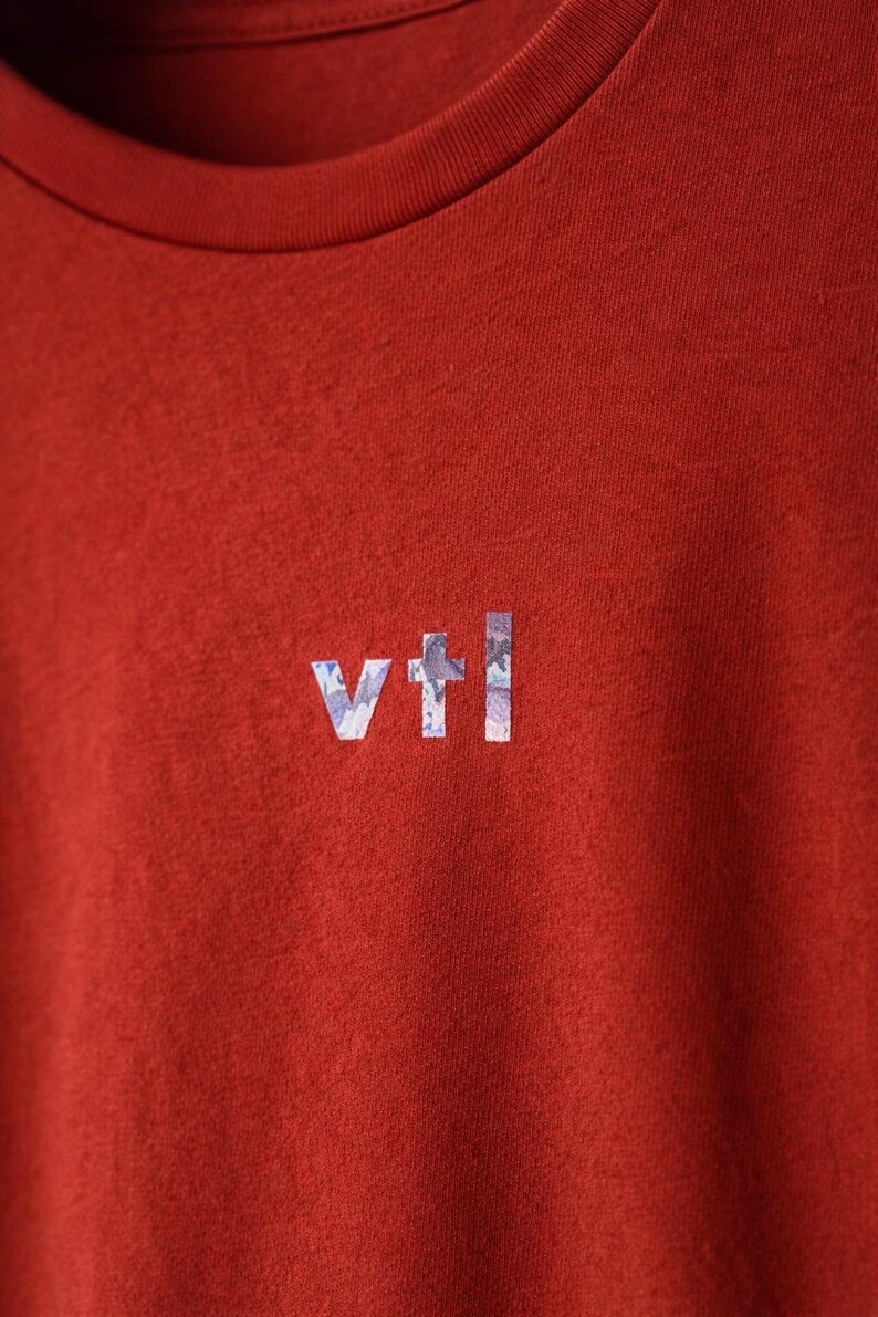May include: Close-up of a rust-colored t-shirt with a crew neck. The shirt features the letters "vtl" in a light blue and white floral pattern on the chest. The fabric has a slightly textured appearance.