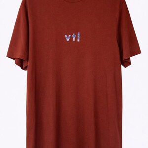May include: A rust-colored t-shirt with short sleeves hanging on a wooden hanger. The shirt features the word "vtl" in a stylized, multi-colored font across the chest. The word "vtl" is also printed on the collar.