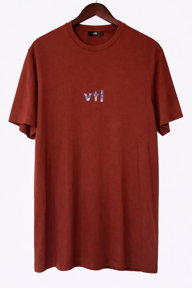 May include: A rust-colored t-shirt with short sleeves hanging on a wooden hanger. The shirt features the word "vtl" in a stylized, multi-colored font across the chest. The word "vtl" is also printed on the collar.