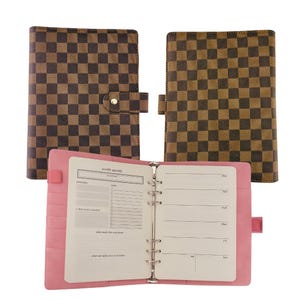 May include: Three brown and black checkered pattern binders, one open to reveal a pink interior with a weekly agenda layout. The binders have a snap closure and a ring binder mechanism.