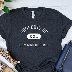 Könnte beinhalten: Dunkelgraues T-Shirt mit dem weißen Text "PROPERTY OF XXL COMMANDER RIP". Das Shirt ist unten geknotet. Leopardenmuster-Sneakers und eine marineblaue Handtasche sind ebenfalls im Bild.