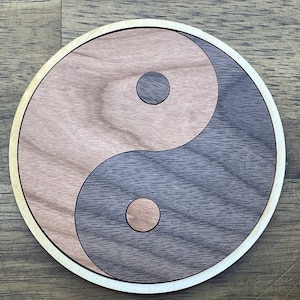 May include: A wooden coaster with a yin yang symbol design. The yin yang symbol is made of two different types of wood, one light brown and one dark brown.