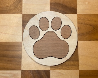 May include: A wooden coaster with a paw print design. The paw print is made of a darker wood and is surrounded by a lighter wood.