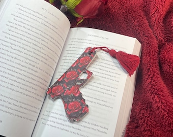 May include: A bookmark shaped like a handgun, covered in red roses, rests on an open book. The bookmark has a red tassel. A red rose and a burgundy blanket are in the background. The book is titled "Haunting Adeline".